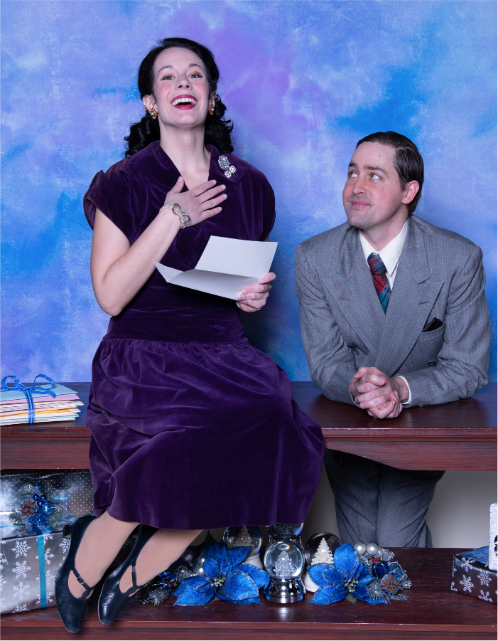 A smiling young woman wearing jewels in a purple velvet dress sits on a bench, a letter in one hand, the other hand to her heart, while a young man in a gray suit sits behind the bench looking lovingly at her.