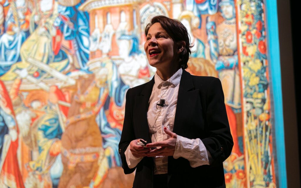 A woman dark jacket and white blouse speaks before a large abstract painting