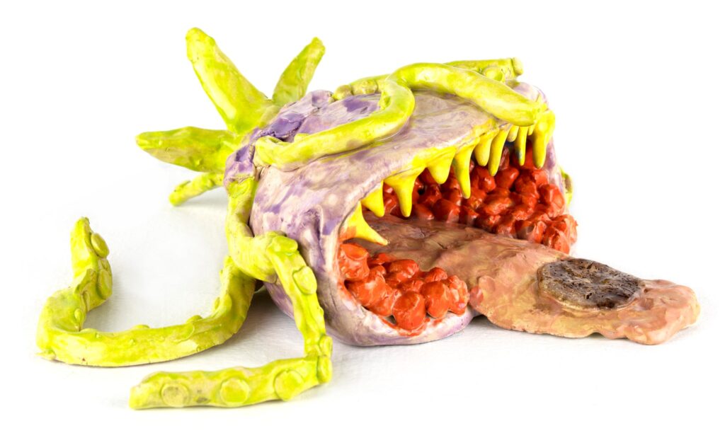 A ceramic sculpture by Kramer Hegenbarth, titled I Sea You Rock, has a big mouth with pointed yellow teeth. The creature's pink tongue extends from its mouth. Greenish-yellow tendrils curl over its purple body and writhe around at its side.