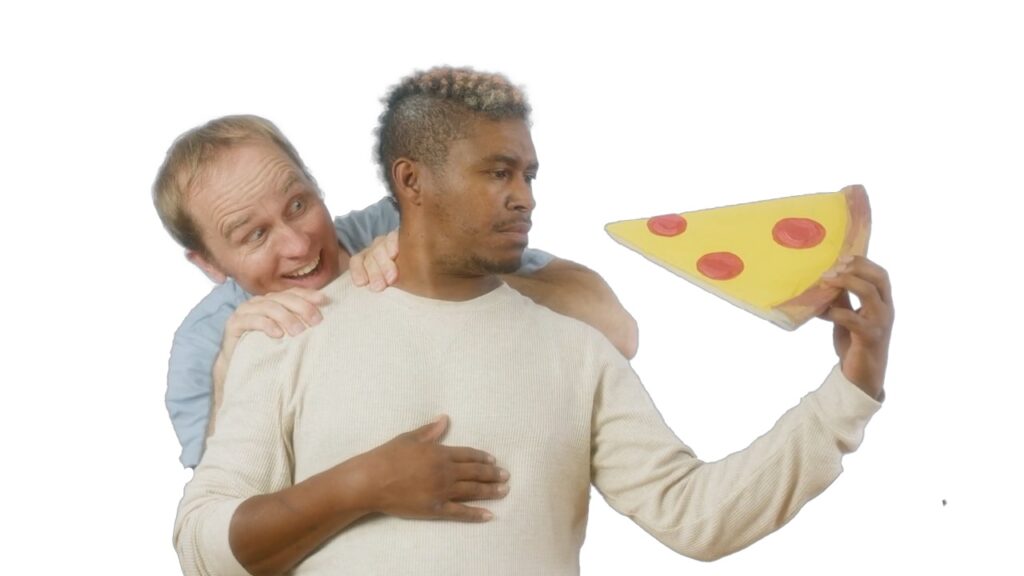 This image shows a man looking at the very large piece of pizza he is holding in his hand. Another man is peaking over his shoulder with a big facial expression of excitement.