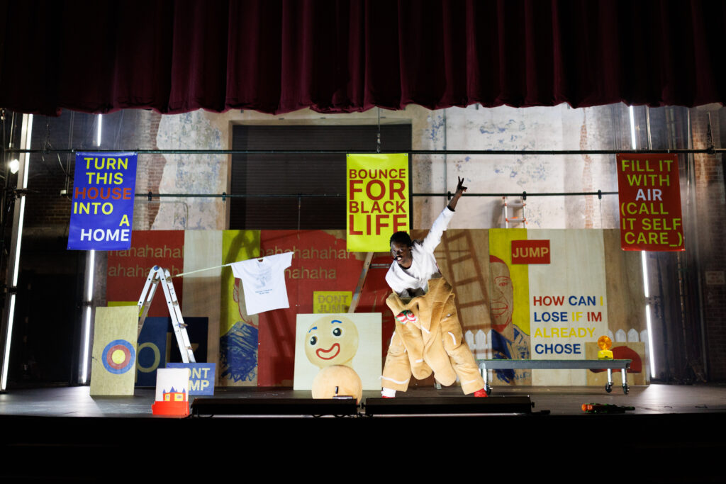 A person in a gingerbread costume poses bent over at the waist with arms out. Behind them is a set of signs with various slogans in blue, red, and yellow. A portrait of George Floyd sits behind them.