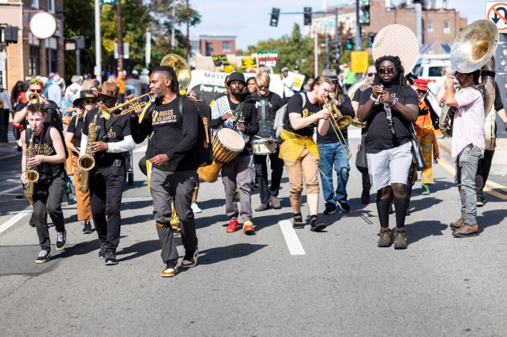 A diverse marching band plays instruments, including brass, drums, and saxophones, while walking down a city street during a daytime parade or protest. People hold signs in the background.