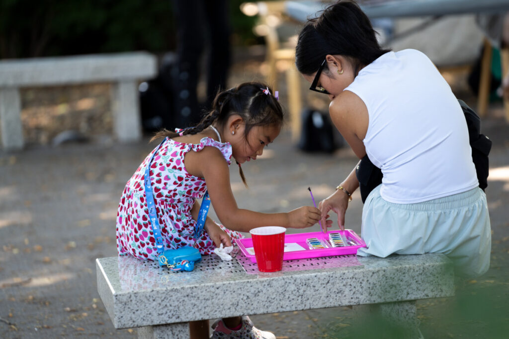 A woman with light skin and dark hair sits to the right of a child with tan skin and dark hair on a grey bench. The child is holding a paint brush and dips it into a watercolor palette.