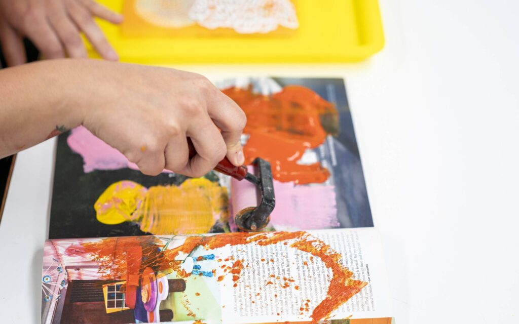 A person uses a paint roller to spread orange paint over a magazine page with colorful images, while a yellow tray with doilies sits nearby on a white surface.