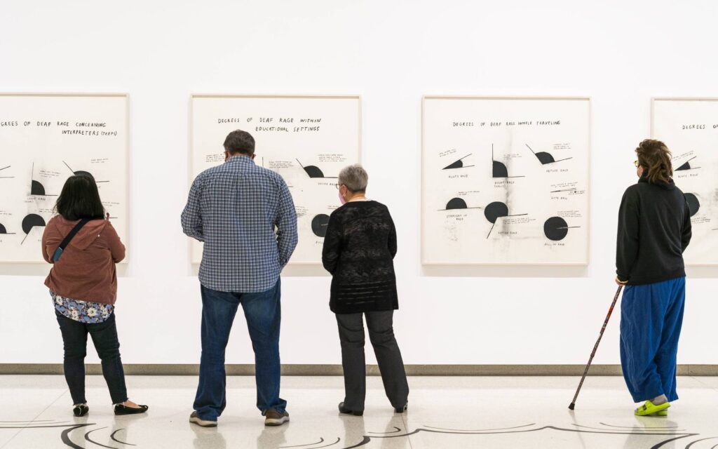 Four people stand in front of white walls, looking at framed black-and-white abstract art with charts and diagrams in a gallery. One person uses a cane. The space is bright and modern.