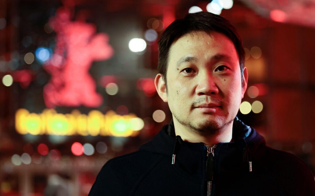 Headshot of Ryusuke Hamaguchi outdoors in a city at night. There are out of focus lights in red and yellow in the background.